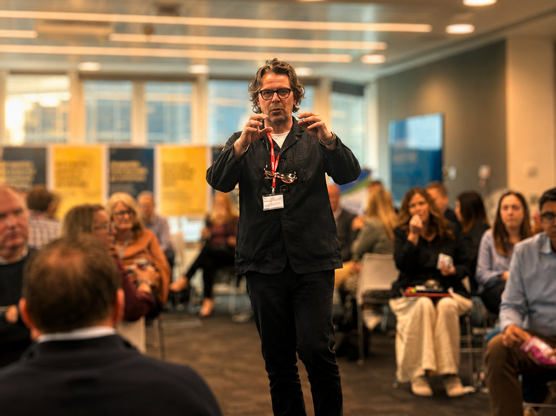 Man speaking to an engaged audience in a modern conference setting, wearing a lanyard and glasses. Attendees sit in chairs, listening attentively.