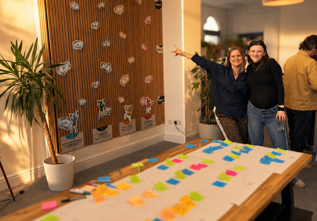 Two people smile and pose beside a colourful board. A table in the foreground is covered with vibrant sticky notes. The mood is joyful and collaborative.